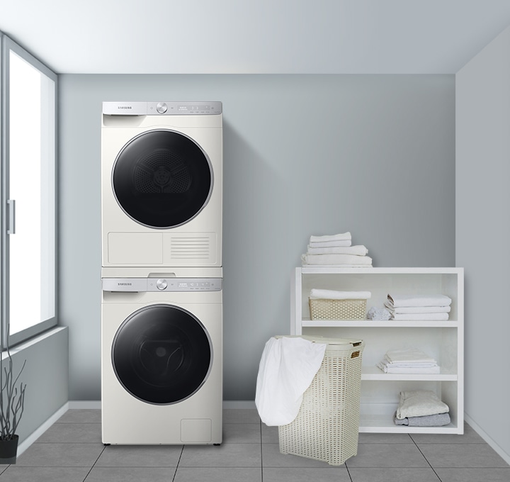 블루톤 벽지의 다용도실 인테리어 컷입니다. 좌측에 창문이 있고 화이트 색상 세탁기/건조기가 직렬 설치되어있습니다. 세탁기/건조기 우측에는 라탄 바구니와 수납장이 있으며 하얀 수건이 보관되어 있습니다. 이미지 우측에는 액자가 걸려있는 벽면입니다.