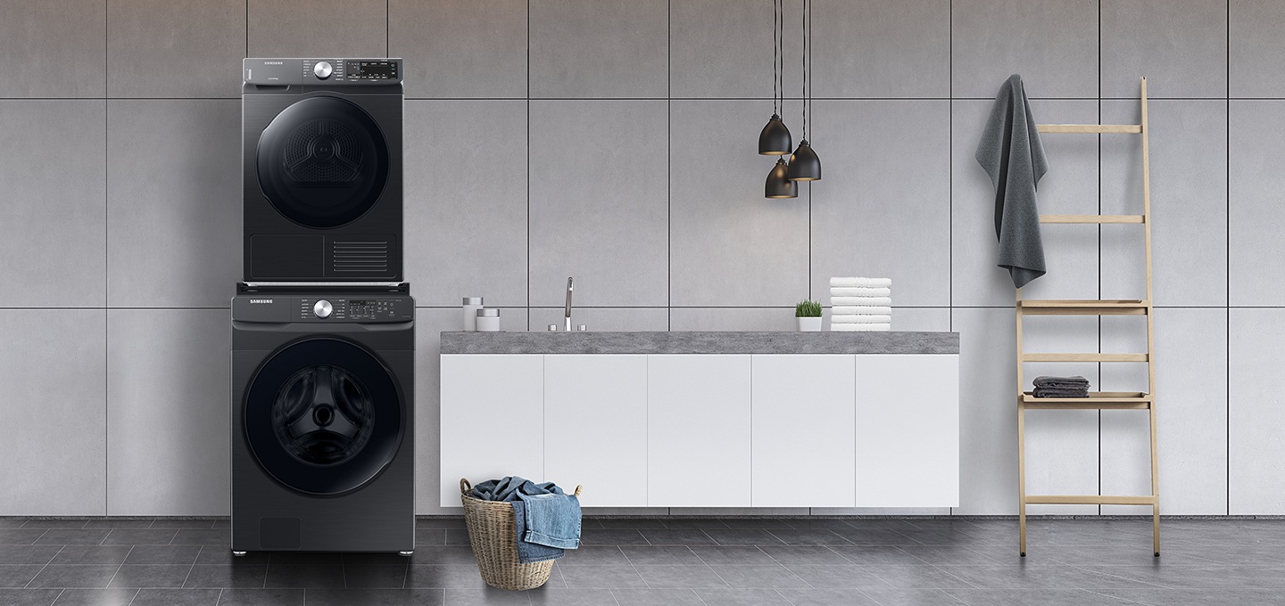 그레이 타일로 인테리어 되어있는 넓은 다용도실이 보이며, 좌측엔 비스포크 그랑데 AI 세탁기 건조기가 건조기 상단키트를 사용해 하나로 되어 있는 모습이 보이며, 가운데에 수납공간이 하단에 함께있는 세면대가 보이며 세면대 위 좌측엔 세탁세제 통이 보이고 우측엔 개어져 있는 하얀색 수건 6개가 보여지고 수건 우측엔 작은 흰 화분이 보여집니다. 세면대 앞엔 라탄의 빨래통과 안에 청바지 빨래가 들어있으며, 우측엔 사다리가 있으며 사딜에 상단에 수건이 걸려있고 두번째 칸에 수건이 개어져 있습니다.