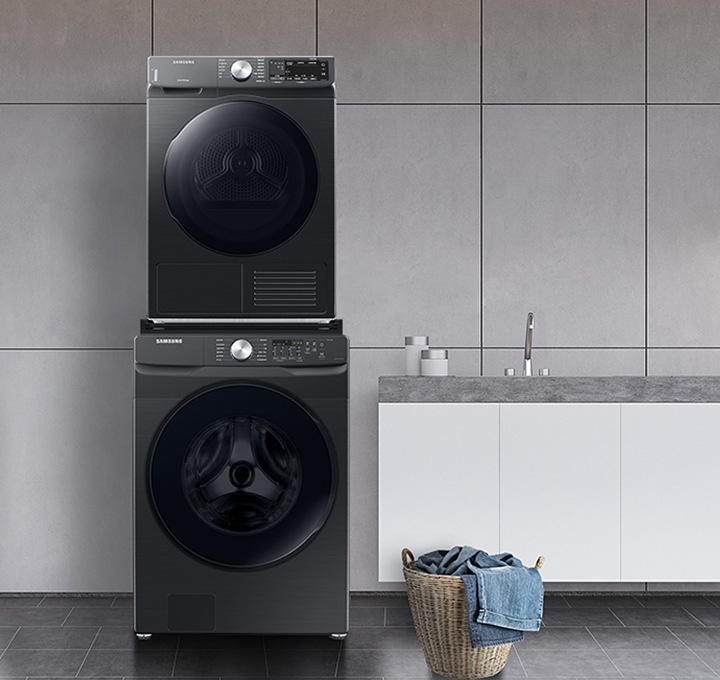 [그레이 타일로 인테리어 되어있는 넓은 다용도실이 보이며, 좌측엔 비스포크 그랑데 ÁÍ 세탁기 건조기가 건조기 상단키트를 사용해 하나로 되어 있는 모습이 보이며, 가운데에 수납공간이 하단에 함께있는 세면대가 보이며 세면대 위 좌측엔 세탁세제 통이 보이고 우측엔 개어져 있는 하얀색 수건 6개가 보여지고 수건 우측엔 작은 흰 화분이 보여집니다. 세면대 앞엔 라탄의 빨래통과 안에 청바지 빨래가 들어있으며, 우측엔 사다리가 있으며 사딜에 상단에 수건이 걸려있고 두번째 칸에 수건이 개어져 있습니다.]