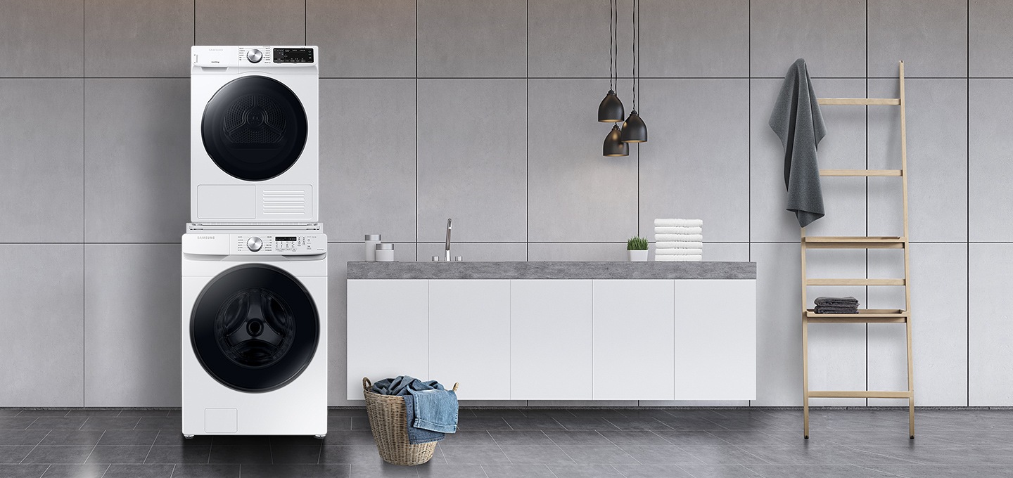 그레이 타일로 인테리어 되어있는 넓은 다용도실이 보이며, 좌측엔 비스포크 그랑데 AI 세탁기 건조기가 건조기 상단키트를 사용해 하나로 되어 있는 모습이 보이며, 가운데에 수납공간이 하단에 함께있는 세면대가 보이며 세면대 위 좌측엔 세탁세제 통이 보이고 우측엔 개어져 있는 하얀색 수건 6개가 보여지고 수건 우측엔 작은 흰 화분이 보여집니다. 세면대 앞엔 라탄의 빨래통과 안에 청바지 빨래가 들어있으며, 우측엔 사다리가 있으며 사딜에 상단에 수건이 걸려있고 두번째 칸에 수건이 개어져 있습니다.