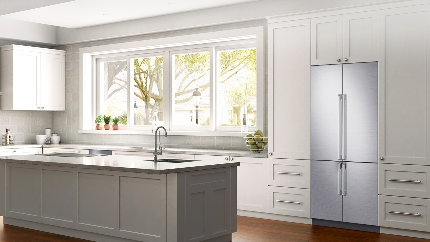 우측면에서 바라본 화이트톤 주방 인테리어컷입니다. 좌측에 아일랜드 식탁이 있고 싱크대 위에 큰 창문이 있습니다. 오른쪽 화이트 냉장고장에 셰프컬렉션 빌트인 모델이 설치되어 있습니다.