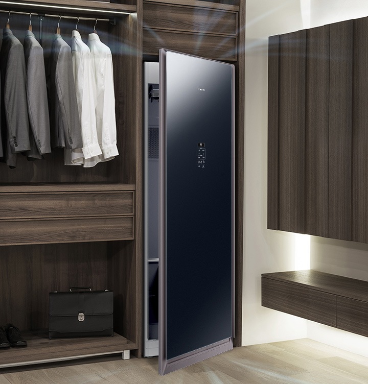 우드톤의 행거옷장이 좌측에 있고 옷장 위쪽에는 와이셔츠와 정장 상의가 여러 벌 걸려있습니다. 행거옷장 중간에는 서랍이 있고 하단에는 블랙 톤의 서류가방이 놓여 있습니다. 바로 옆 우측에는 에어드레서 드레스룸을 제습하고 있는 모습입니다.