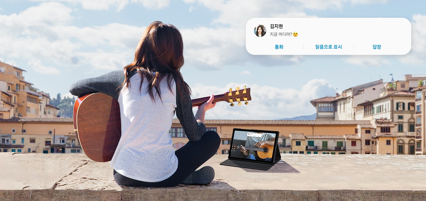 한 여성이 옥상 바닥에 앉아 태블릿으로  영상을 보며 기타를 치고 있습니다. 태블릿 화면 상단에는  김지현이라는 친구에게온 문자 알람이 떠있습니다. 문자에는 지금 어디야? 라고 쓰여져 있습니다.