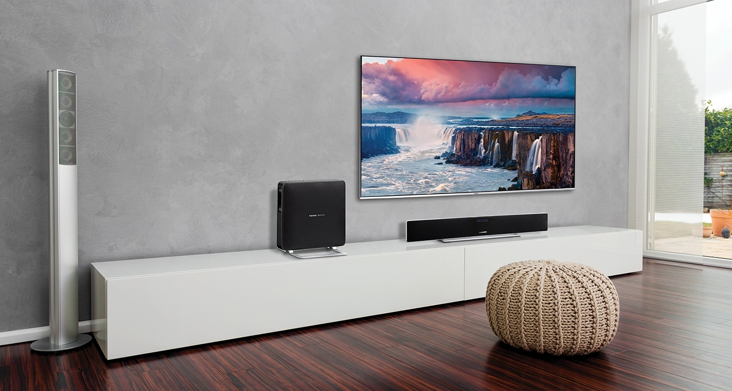 거실에 벽걸이형 TV가 걸려 있습니다. TV 앞과 옆에는 사운드바와 우퍼가 놓여져 있습니다.