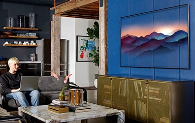 거실 벽면에 QLED TV가 켜진 상태로 걸려있고 쇼파에 앉은 사람이 노트북을 사용하고 있습니다. 