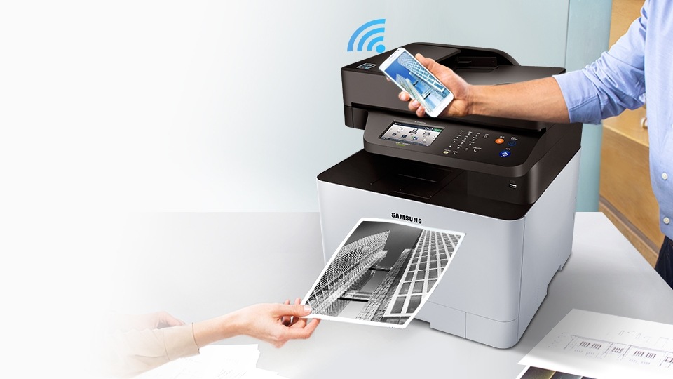 탁자위에 SL-C1453FW 모델이 있고 뒤에는 노트북이 있으며 출력물을 들고 있는 손과 스마트폰을 들고 있는 손이 연출됨