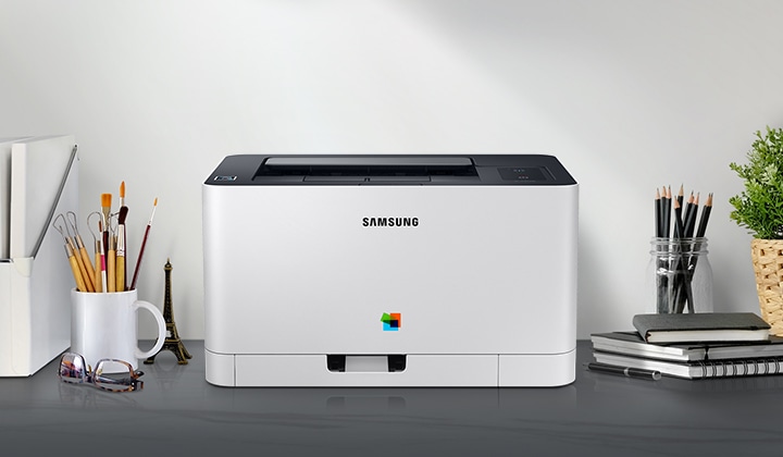 컬러 레이저프린터 18/4 ppm 화이트 제품 정면이 그레이 색상의 책상 위에 올려져 있고 주변에 사무용품들이 배치되어 있습니다.