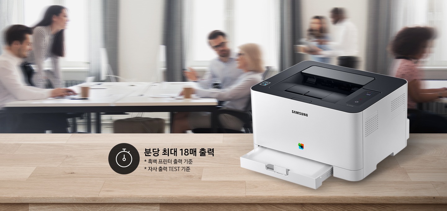 컬러 레이저프린터 18/4 ppm 화이트 제품이 측면으로 우드 책상 위에 올려져 있고, 그 뒤에는 회의를 하는듯한 배경 이미지가 블러처리 되어 있습니다.