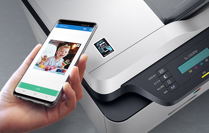 검정 색상의 나무 책상에 제품의 왼쪽 위에서 내려본 이미지가 있고 스마트폰을 제품 이미지 상단에 NFC 태그 스티커가 붙어있는 부분에 태그하는 이미지입니다.