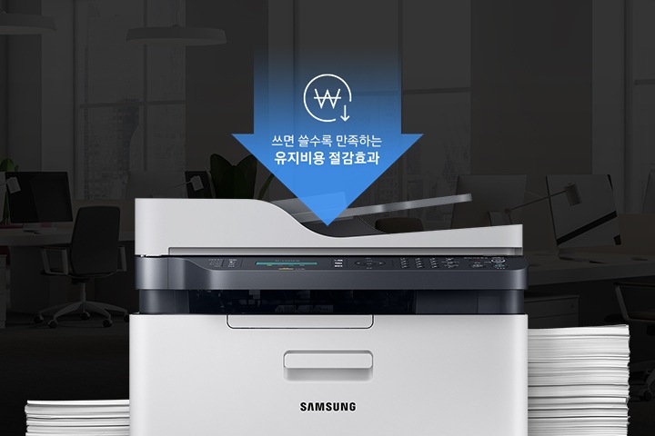 어두운 사무실 배경에 이미지 중간에는 제품 정면 이미지가 놓여있고 제품 이미지 양옆으로는 종이가 쌓여있습니다. 제품 이미지 상단에는 아래로 내려오는 파란 색상에 화살표와 화살표 안에 쓰면 쓸수록 만족하는 유지비용 절감효과 텍스트와 동그라미 안에 원 표시와 오른쪽 하단에 아래로 내려가는 아이콘이 있습니다.