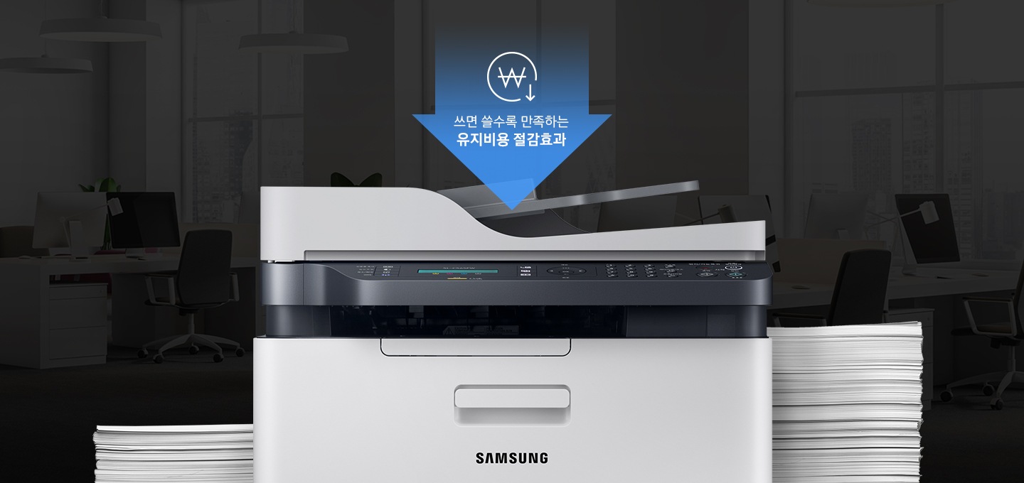 어두운 사무실 배경에 이미지 중간에는 제품 정면 이미지가 놓여있고 제품 이미지 양옆으로는 종이가 쌓여있습니다. 제품 이미지 상단에는 아래로 내려오는 파란 색상에 화살표와 화살표 안에 쓰면 쓸수록 만족하는 유지비용 절감효과 텍스트와 동그라미 안에 원 표시와 오른쪽 하단에 아래로 내려가는 아이콘이 있습니다.