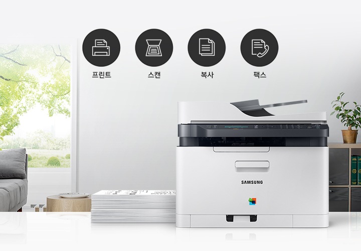 이미지 왼쪽에는 푸른 나무가 보이는 창문과 회색 소파가 배치되어 있으며, 이미지 오른쪽에는 제품 정면 이미지가 배치되어 있습니다. 제품 이미지 왼쪽에는 종이가 쌓여 있으며, 제품 이미지와 소파 사이에는 프린터 텍스트와 프린터 아이콘, 스캔 텍스트와 스캔 아이콘, 복사 텍스트와 복사 아이콘, 팩스 텍스트와 팩스 아이콘이 왼쪽에서 오른쪽으로 나열하여 있습니다.