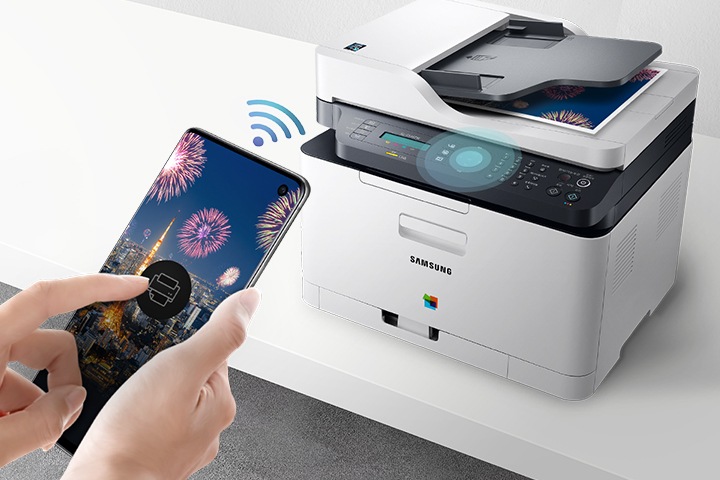 하얀색 책상 위에 제품을 오른쪽 위에서 내려본 이미지가 중간에 배치하여 있고 이미지 왼쪽 부분에 스마트폰 화면에 프린터 아이콘을 누르고 있는 사람의 손이 배치하여 있습니다.