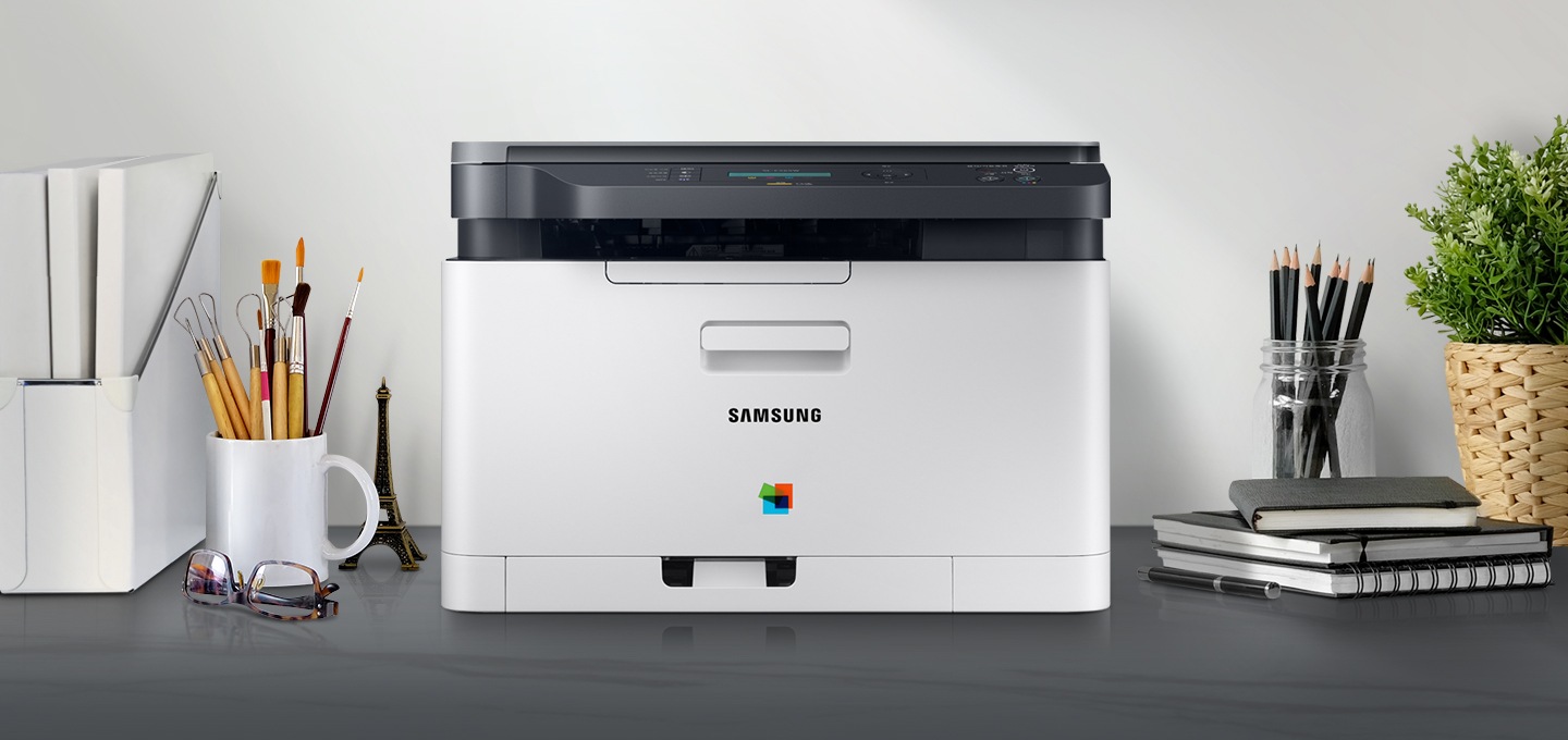 컬러 레이저복합기 18/4 ppm 화이트 제품 정면이 그레이 색상의 책상 위에 올려져 있고 주변에 사무용품들이 배치되어 있습니다.