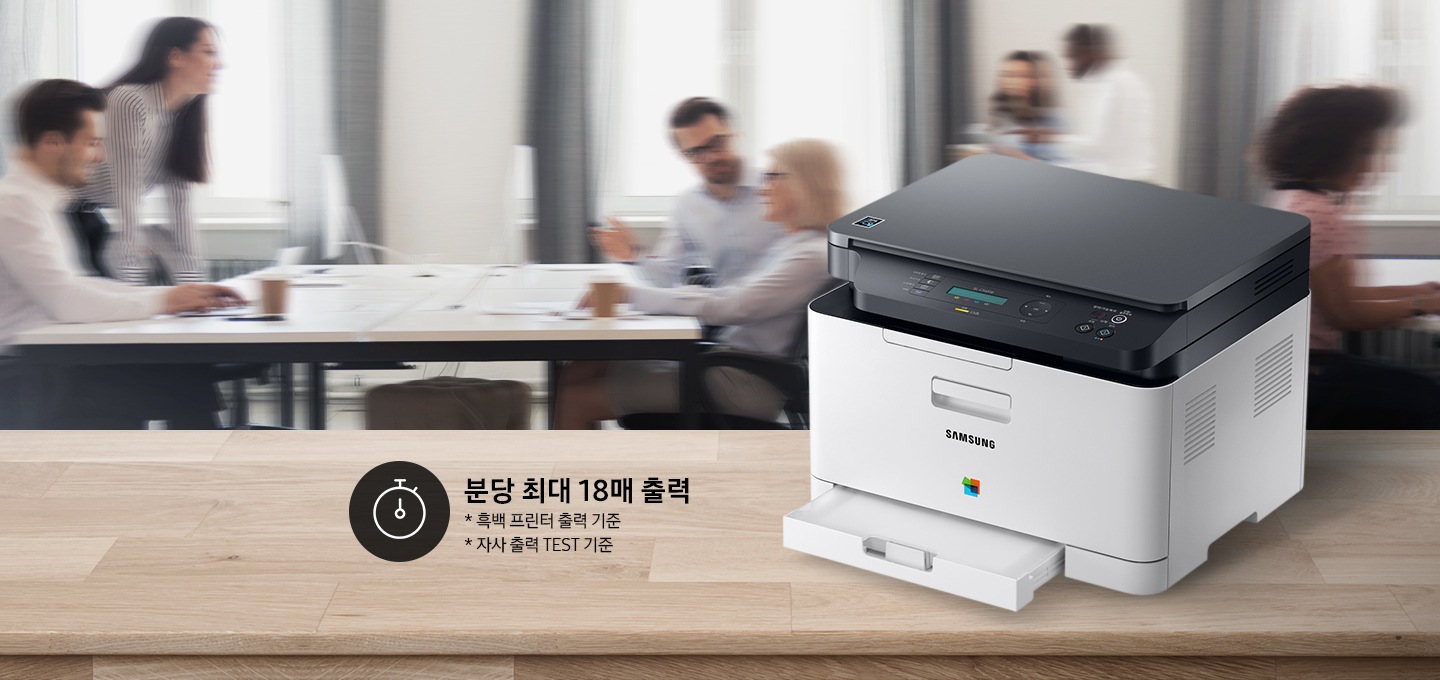 컬러 레이저복합기 18/4 ppm 화이트 제품이 측면으로 우드 책상 위에 올려져 있고, 그 뒤에는 회의를 하는듯한 배경 이미지가 블러처리 되어 있습니다.