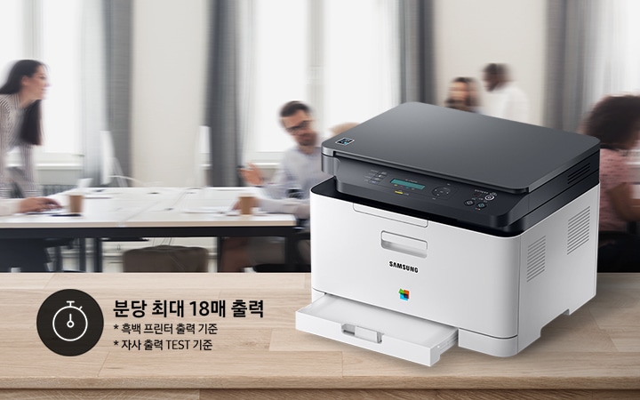 컬러 레이저복합기 18/4 ppm 화이트 제품이 측면으로 우드 책상 위에 올려져 있고, 그 뒤에는 회의를 하는듯한 배경 이미지가 블러처리 되어 있습니다.