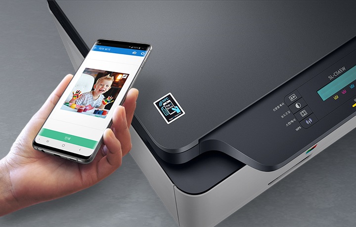 프린터에 NFC를 연결하여 스마트폰의 이미지를 출력 할 수 있음을 알려주는 이미지 입니다. 프린터 NFC라고 적힌 아이콘 위에 스마트폰을 들고 있고, 스마트폰 배경에는 아이의 사진이 있습니다.