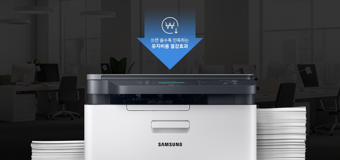 어두운 사무실 배경에 이미지 중간에는 제품 정면 이미지가 놓여있고 제품 이미지 양옆으로는 종이가 쌓여있습니다. 제품 이미지 상단에는 아래로 내려오는 파란 색상에 화살표와 화살표 안에 쓰면 쓸수록 만족하는 유지비용 절감효과 텍스트와 동그라미 안에 원 표시와 오른쪽 하단에 아래로 내려가는 아이콘이 있습니다.