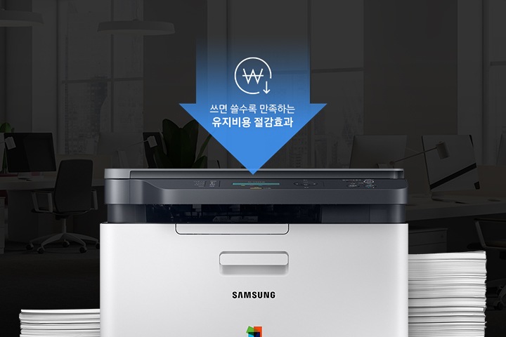 어두운 사무실 배경에 이미지 중간에는 제품 정면 이미지가 놓여있고 제품 이미지 양옆으로는 종이가 쌓여있습니다. 제품 이미지 상단에는 아래로 내려오는 파란 색상에 화살표와 화살표 안에 쓰면 쓸수록 만족하는 유지비용 절감효과 텍스트와 동그라미 안에 원 표시와 오른쪽 하단에 아래로 내려가는 아이콘이 있습니다.