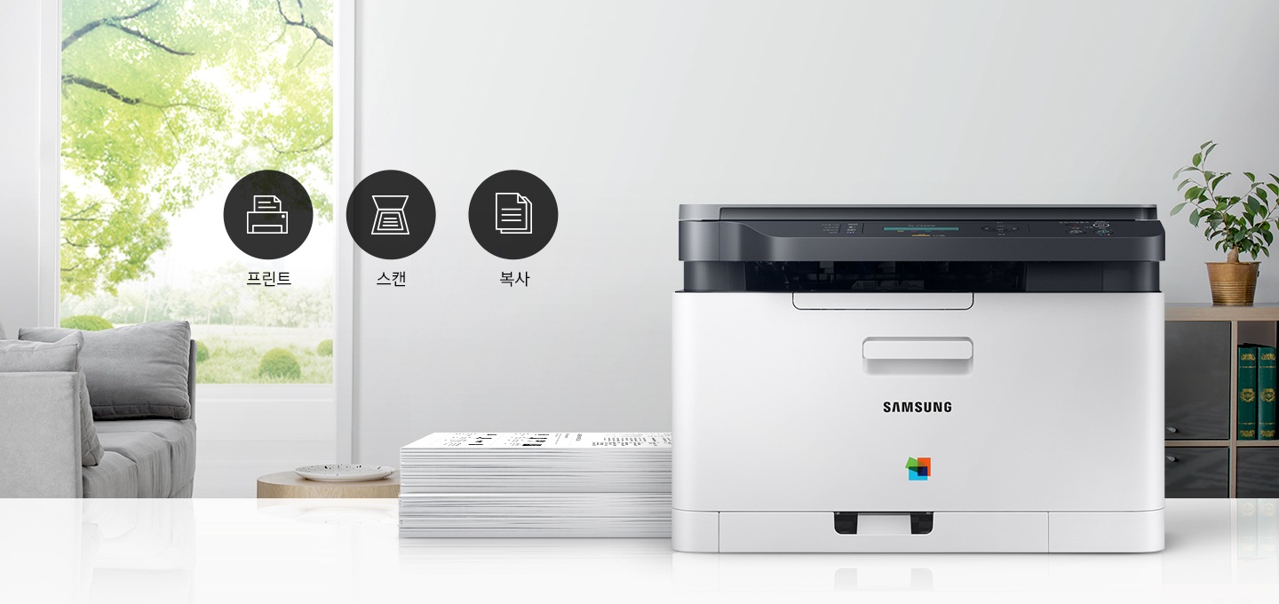 이미지 왼쪽에는 푸른 나무가 보이는 창문과 회색 소파가 배치되어 있으며, 이미지 오른쪽에는 제품 정면 이미지가 배치되어 있습니다. 제품 이미지 왼쪽에는 종이가 쌓여 있으며, 제품 이미지와 소파 사이에는 프린터 텍스트와 프린터 아이콘, 스캔 텍스트와 스캔 아이콘, 복사 텍스트와 복사 아이콘이 왼쪽에서 오른쪽으로 나열하여 있습니다.
