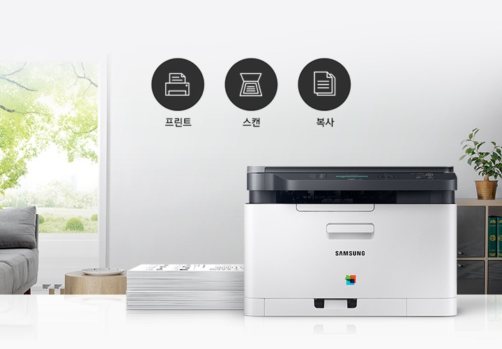 이미지 왼쪽에는 푸른 나무가 보이는 창문과 회색 소파가 배치되어 있으며, 이미지 오른쪽에는 제품 정면 이미지가 배치되어 있습니다. 제품 이미지 왼쪽에는 종이가 쌓여 있으며, 제품 이미지와 소파 사이에는 프린터 텍스트와 프린터 아이콘, 스캔 텍스트와 스캔 아이콘, 복사 텍스트와 복사 아이콘이 왼쪽에서 오른쪽으로 나열하여 있습니다.