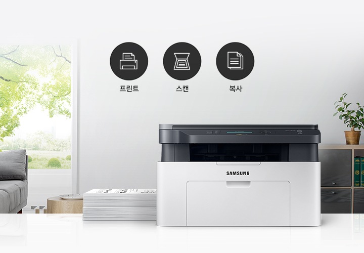 이미지 왼쪽에는 푸른 나무가 보이는 창문과 회색 소파가 배치되어 있으며, 이미지 오른쪽에는 제품 정면 이미지가 배치되어 있습니다. 제품 이미지 왼쪽에는 종이가 쌓여 있으며, 제품 이미지와 소파 사이에는 프린터 텍스트와 프린터 아이콘, 스캔 텍스트와 스캔 아이콘, 복사 텍스트와 복사 아이콘이 왼쪽에서 오른쪽으로 나열하여 있습니다.