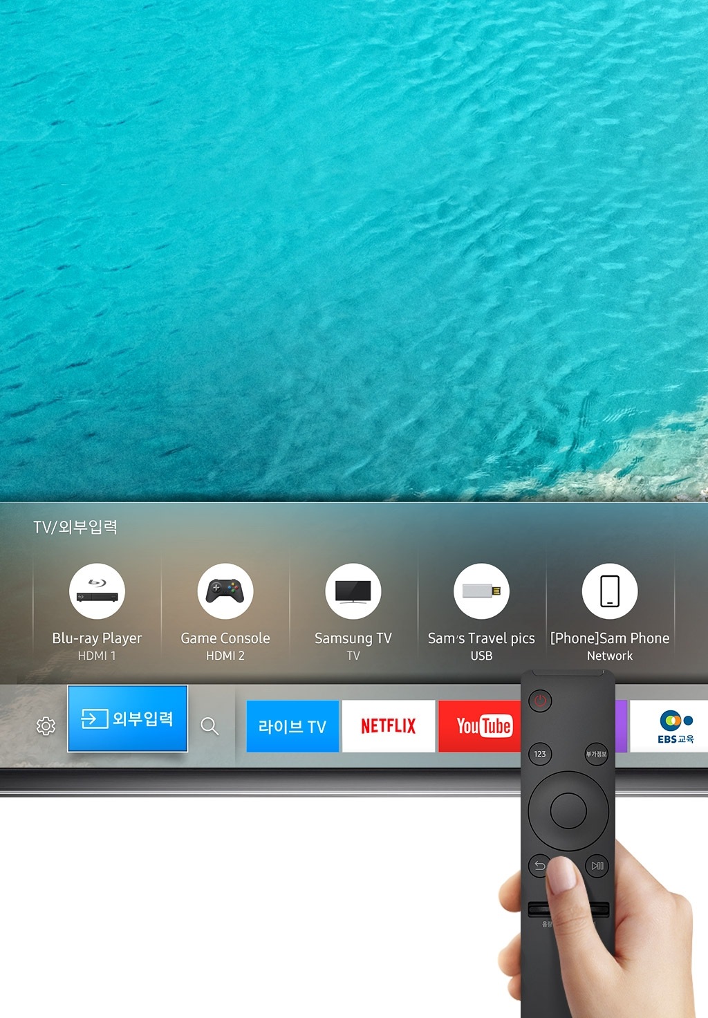 화면의 상단에 삼성 UHD TV가 확대되어 나타나 있습니다. 제품은 하단부분만 확대되어 나타나 있으며, 해변배경의 화면 위로 TV/외부입력, 콘텐츠들이 bar 형태의 메뉴로 나타나 있습니다. 그런 제품의 위로 오른손으로 검정 색상의 TV리모컨을 쥐고 있는 모습이 보여집니다.
