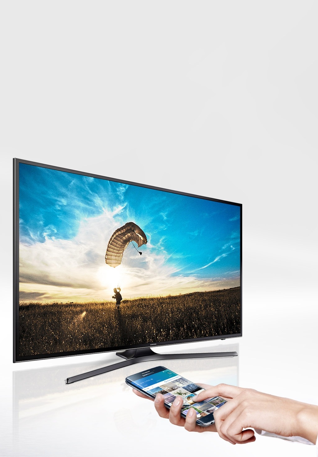 회색 빛의 화면의 좌측에 삼성 UHD TV가 대각선으로 비스듬히 위치해 있습니다. 제품의 화면에는 갈색 평원에 패러글라이딩을 하며 착륙하는 남자가 보입니다. 남자의 뒤로 태양이 밝게 비춰지며, 구름낀 파란하늘이 보여집니다. 그런 제품의 오른쪽 하단에는 흰색 소매의 옷을 입은 여성의 손이 오른손으로는 스마트폰을 쥐고 왼손으로는 사진을 선택하고 있습니다. 왼손의 검지가 가리키는 스마트폰의 사진과 삼성 UHD TV의 사진은 동일한 사진으로 나타납니다.