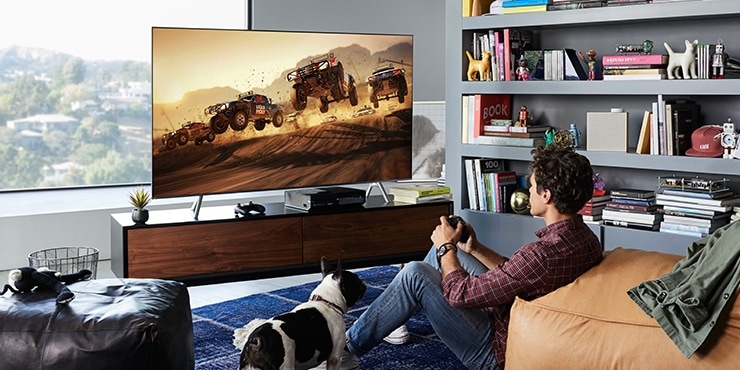 선반 위에 QLED TV가 정면으로 놓여져 있으며, 화면 안에는 게임의 한 장면이 보여집니다. 그 앞에 한 청년이 소파에 기대어 앉아 게임을 즐기고 있습니다.