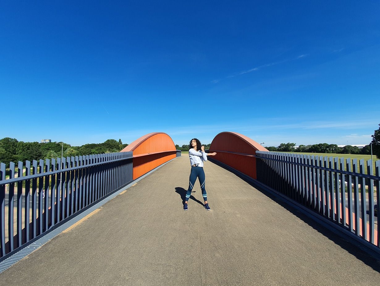 Foto di una donna che fa ginnastica su un ponte dalla struttura grigia e note arancio sullo sfondo di un cielo blu acceso realizzata con la fotocamera ultra-grandangolare