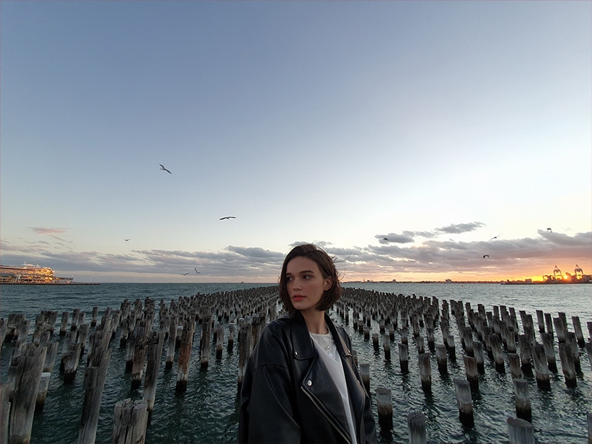 Foto di una donna al tramonto in piedi davanti a dei pali di legno che si ergono dal mare, scattata con Galaxy S10 Plus. Mostra una visione piÃ¹ ampia della scena, grazie alla fotocamera ultra-grandangolare.
