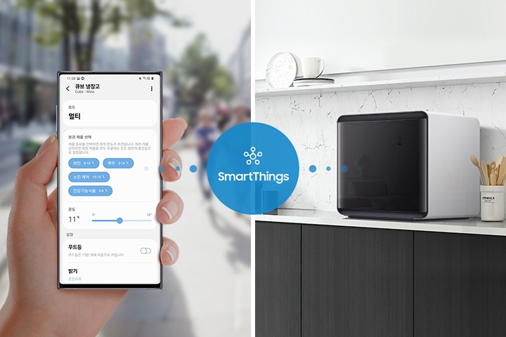 좌측에는 휴대폰을 쥐고 있는 손 사진이 나와있습니다. 휴대폰 화면애는 SmartThings 큐브 냉장고 화면이 나와있으며 모드와 온도, 보관 제품 선택 등의 옵션이 보여집니다. 가운데 SmartThings  로고와 문구가 파란 원 안에 있으며 우측 화이트톤 인테리어컷 선반 위에 설치된 BESPOKE 큐브 냉장고와 연결되고 있는 이미지입니다.
