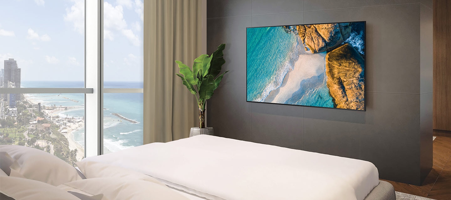 창밖으로 바다가 보이는 호텔방이 있으며, 침대 정면 벽에는 호텔TV CU700 시리즈가 설치된 모습입니다.