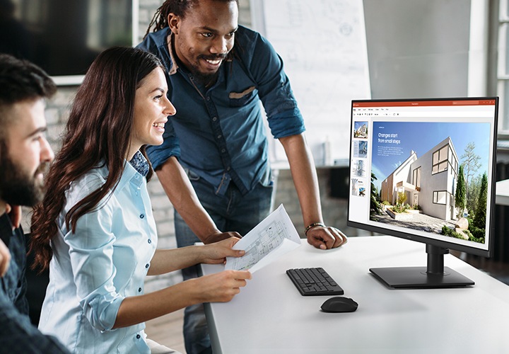 책상 위에 키보드 및 마우스와 모니터가 보입니다. 모니터 화면에는 건물이 잘 보이는 풍경이 보입니다. 모니터 앞에는 여성이 설계도를 양 손에 쥔 채 모니터를 웃으며 바라보고 있습니다. 이 여성 주위로 남성 2명과 여성 1명이 함께 모니터를 바라보고 있습니다.