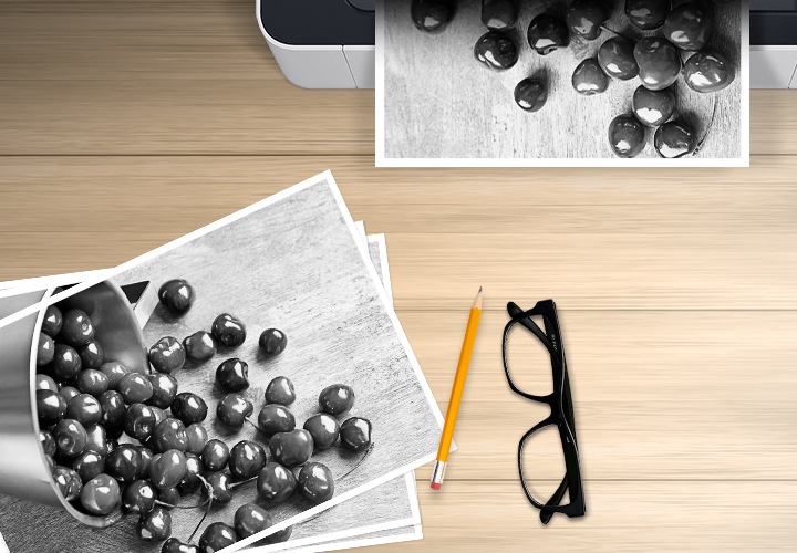 나무로된 책상 배경에 이미지 왼쪽 하단 부분에는 체리가 쏟아진 이미지를 출력한 출력물이 있고 이미지 오른쪽 상단에는 체리가 쏟아진 이미지를 출력중인 프린터를 위에서 내려본 이미지다 있습니다. 이미지 오른쪽 하단에는 연필과 안경 이미지가 보여지고 있습니다.