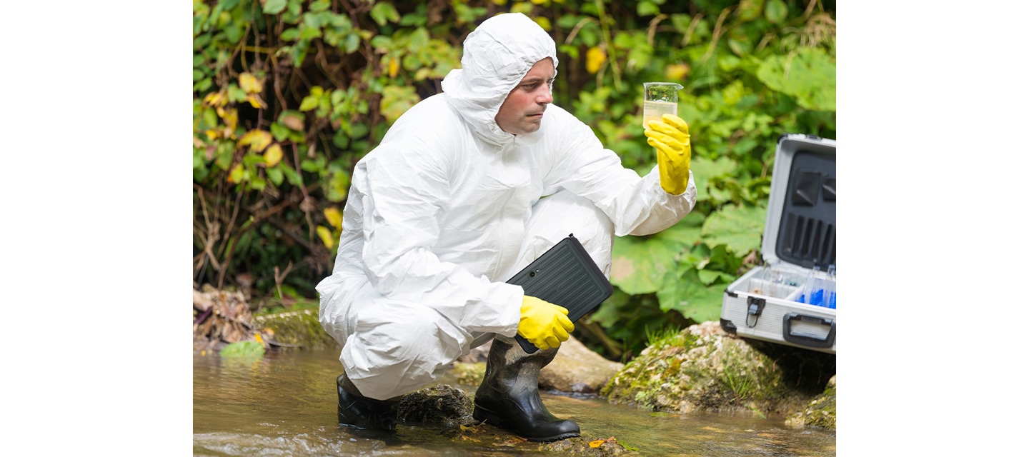 방진복과 보호 장비를 착용한 남성이 호숫가에서 수질 샘플을 채취하고 있습니다. 왼손에는 채취한 물이 담긴 비커, 오른손에는 갤럭시 탭 액티브5 프로를 들고 쪼그려앉아 있습니다. 기기가 물, 먼지 및 오염물에 강한 저항성을 지니고 있음을 명확하게 표현하고 있습니다.