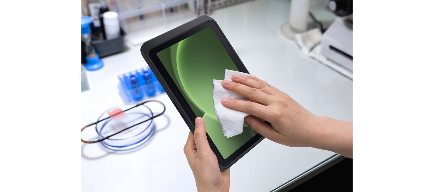 실험실 환경에서 갤럭시 탭 액티브5 프로를 세척하는 모습입니다. 왼손은 갤럭시 탭 액티브5 프로를 잡고 있으며 오른손에는 천을 들고 화면을 닦고 있습니다. 디스플레이에는 녹색 음영의 그래픽이 띄워져 있습니다.