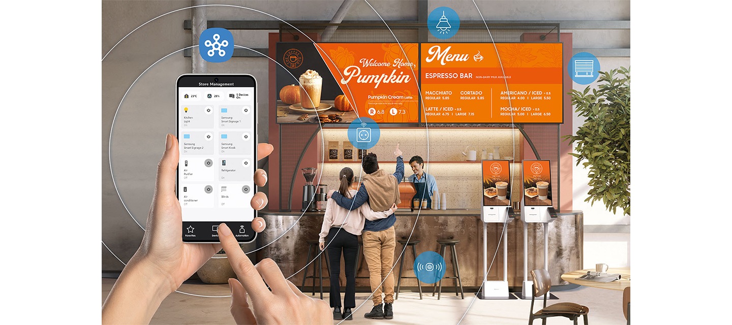 벽면에 비즈니스TV가 설치된 카페의 카운터를 바라보고 있으며, 그 앞으로 스마트폰을 들고 있는 손이 보입니다. 스마트폰 화면에는 설치된 비즈니스TV의 화면을 App으로 제어할 수 있는 화면이 보입니다.
