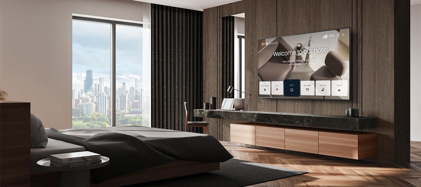 바깥 도시풍경이 보이는 고급스러운 호텔방의 침대 정면에 삼성 호텔TV가 설치되어 있고, 화면으로 호텔의 안내 화면이 송출되고 있는 모습입니다.