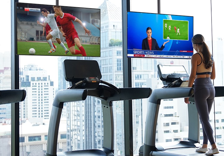 통창으로 바깥 풍경이 보이는 헬스장의 창 앞에 런닝머신이 있으며, 러닝머신의 정면으로 각 축구경기와 뉴스를 송출중인 호텔 TV가 보입니다. 러닝머신 한곳에서는 한 여성이 운동을 하고 있는 모습입니다.