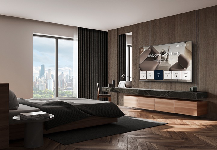 바깥 도시풍경이 보이는 고급스러운 호텔방의 침대 정면에 삼성 호텔TV가 설치되어 있고, 화면으로 호텔의 안내 화면이 송출되고 있는 모습입니다.