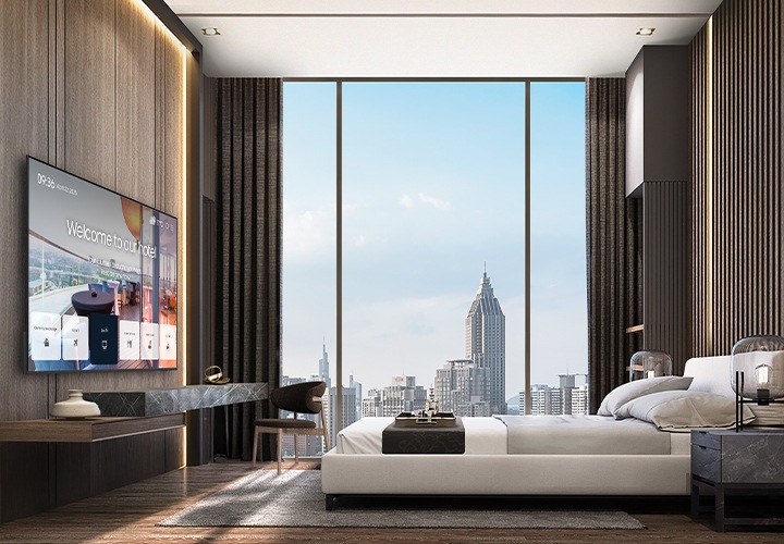 바깥 도시풍경이 보이는 고급스러운 호텔방의 침대 정면에 삼성 호텔TV가 설치되어 있고, 화면으로 호텔의 안내 화면이 송출되고 있는 모습입니다.