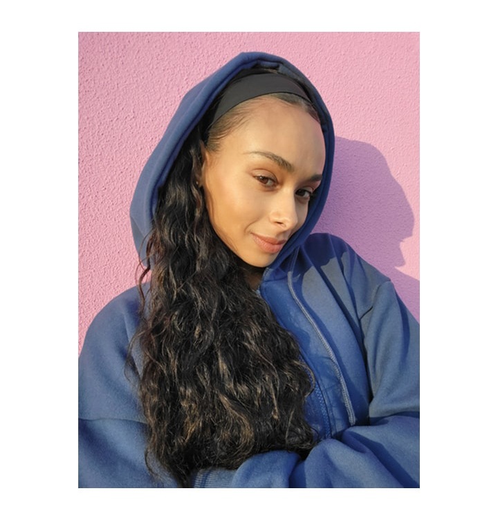 핑크색 벽에 기대어 한 여성이 고개를 비스듬히 한 채 웃으면서 셀카를 찍고 있습니다.