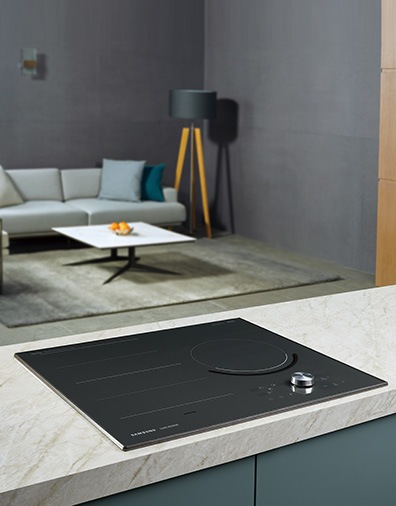 대리석으로 된 조리대 위에 전기레인지가 빌트인 방식으로 설치되어 있으며 뒤로 보이는 거실에는 밝은색의 소파와 사각테이블이 놓여 있습니다.