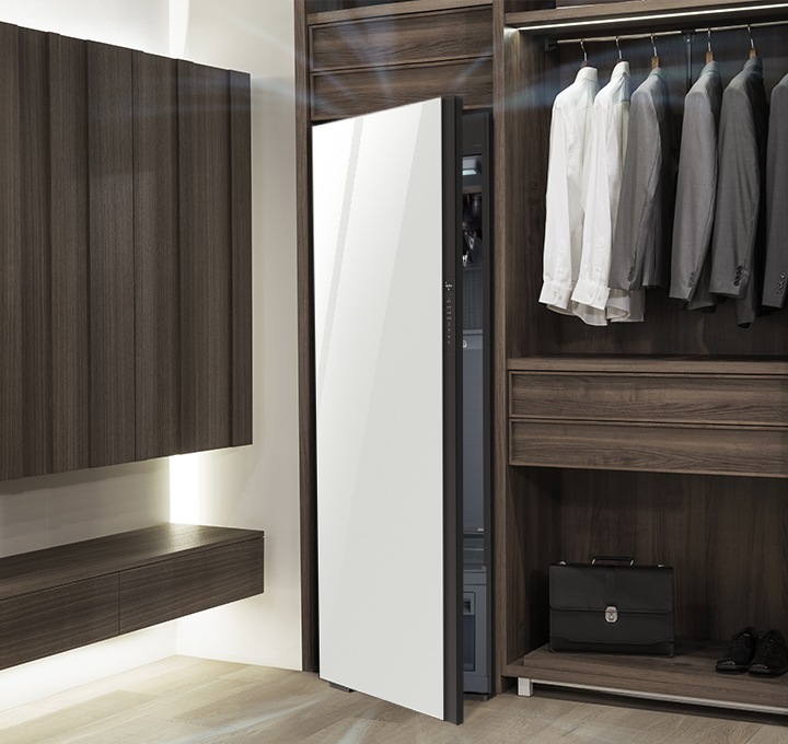 우드톤의 행거옷장이 좌측에 있고 옷장 위쪽에는 와이셔츠와 정장 상의가 여러 벌 걸려있습니다. 행거옷장 중간에는 서랍이 있고 하단에는 블랙 톤의 서류가방이 놓여 있습니다. 바로 옆 우측에는 에어드레서 드레스룸을 제습하고 있는 모습입니다.