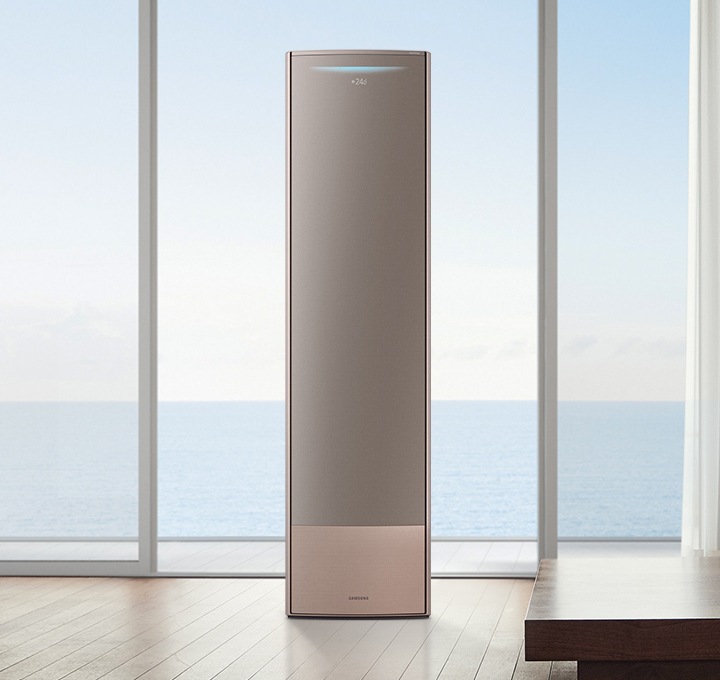 통창 너머로 바다가 보이는 거실에 짙은 다크 브라운의 바닥과 벽면이 보이며 좌식 테이블과 쿠션들이 보입니다.통창의 앞에는 저채도의 골드컬러 패널이 설치된 비스포크 무풍 에어컨 갤러리1가 설치되어 있습니다. 