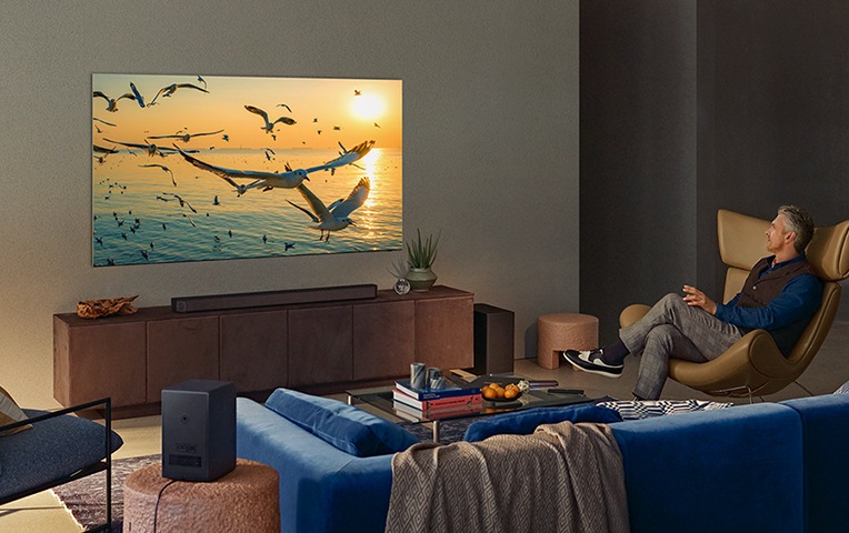남선 사용자가 쇼파에 앉아 TV를 보고 있습니다. TV 에는 해변과 새 이미지가 나오고 있고 하단에는 사운드바가 설치되어 있습니다. 사용자 주변에 서브우퍼도 함께 설치되어있습니다.