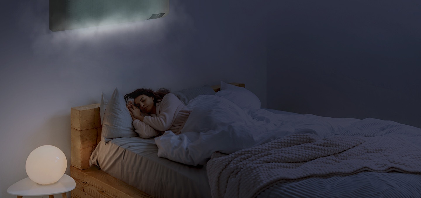 에어컨이 설치된 방에서 여성이 침대에 누워 자고 있는 모습이 보여집니다. 에어컨에서는 은은한 냉기가 뿜어져 나오고 있습니다.