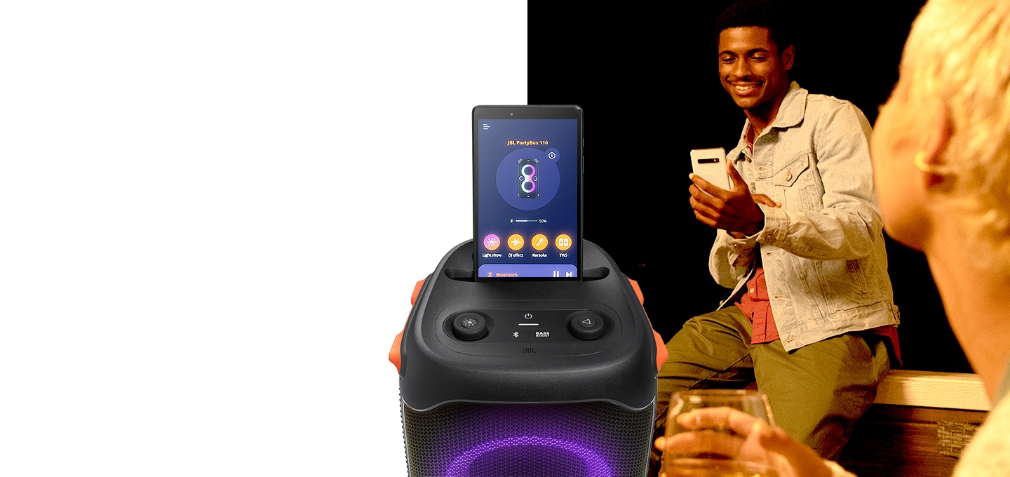 남성 사용자가 스마트폰을 보고 있습니다. 좌측에는 PARTYBOX 110 제품의 정면컷이 보이고 있고 상단에 태블릿이 연결되어 있습니다. 화면에는 PARTYBOX 110 제품의 온스크린이 보입니다.