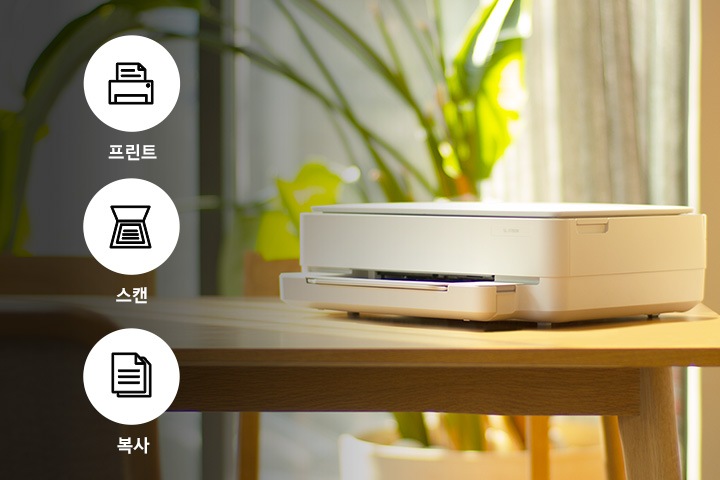 햇빛이 쏟아지는 창문 앞네 식물이 닜는 배경에 식탁 위에 제품이 왼쪽으로 20도 돌아간 이미지가 왼쪽에 배치되어 있는 사진입니다. 제품 이미지 왼쪽에는 프린터 텍스트와 프린터 이미지, 스캔 텍스트와 스캔 이미지, 복사 텍스트와 복사 아이콘이 왼쪽에서 오른쪽으로 나열되어 있는 이미지 입니다.